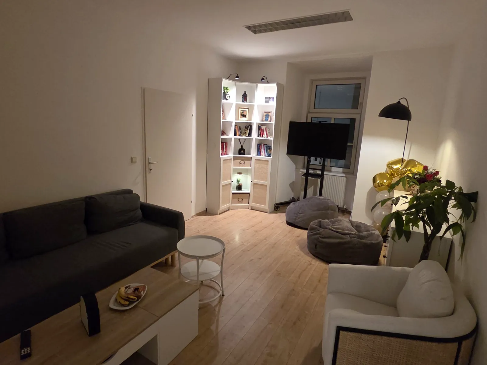 Die Lounge mit dunklem Sofa, weißem Sessel, Sitzsäcken, beleuchtetem Bücherregal und Pflanzen