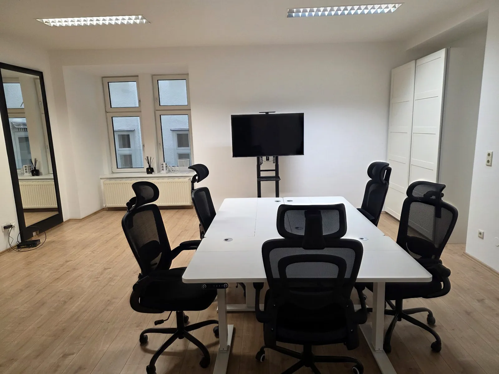 Ein Tisch mit sechs ergonomischen Stühlen vor einem großen Bildschirm, daneben ein hoher Spiegel und zwei Fenster