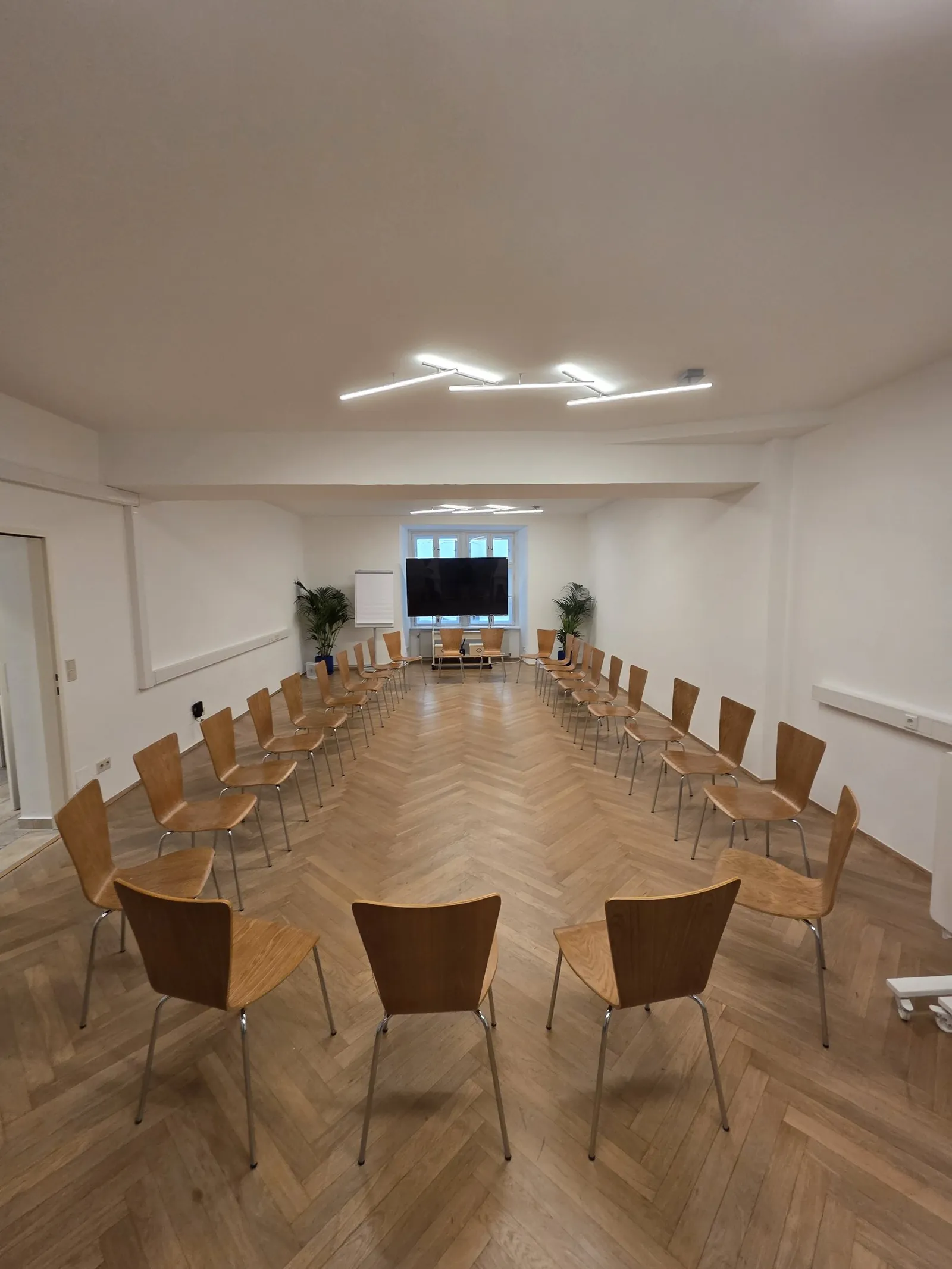 Der Seminarraum mit Holzstühlen im Stuhlkreis, Pflanzen, Flipchart und großem Bildschirm