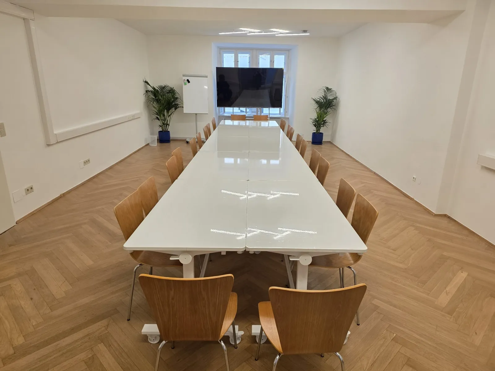 Der Seminarraum mit langem weißem Tisch, Holzstühlen, Pflanzen, Flipchart und großem Bildschirm