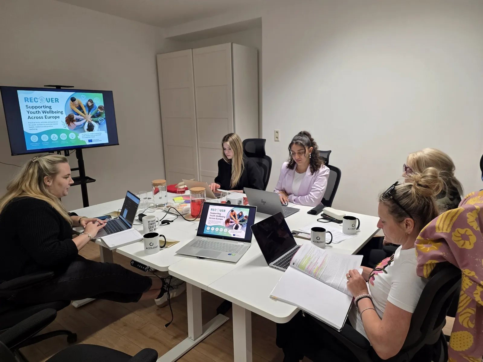 Teammitglieder mit Laptops und ACCELERAS-Tassen an einem Tisch, eine Projektfolie auf dem Bildschirm